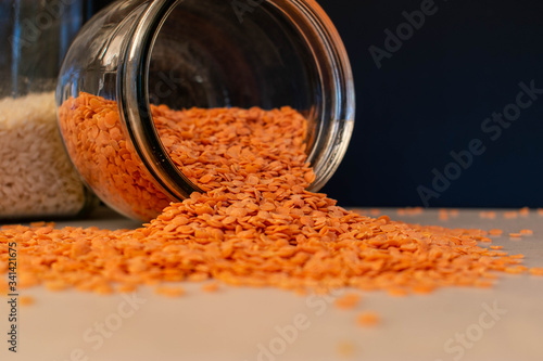 A close up of red lentils spilling out of a glass storage container onto a plain work top against a black background photo