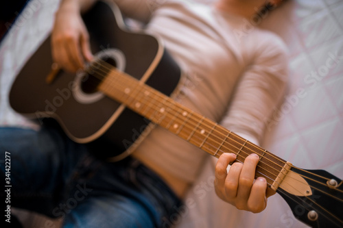 Young man in a light jumper and jeans plays an acoustic guitar in the morning in bed