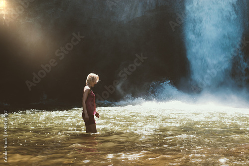 Young blonde woman with pink blouse and shorts walking in brown water river and illuminated by the sunbeam in front of the running blue waterfall in Bali island