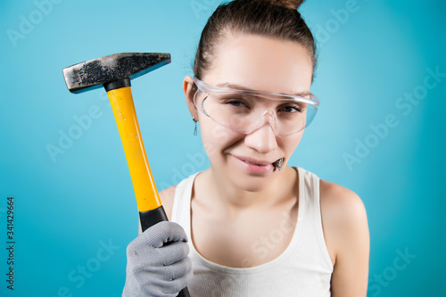 The girl in goggles holds a hammer in her hand and nails in her teeth