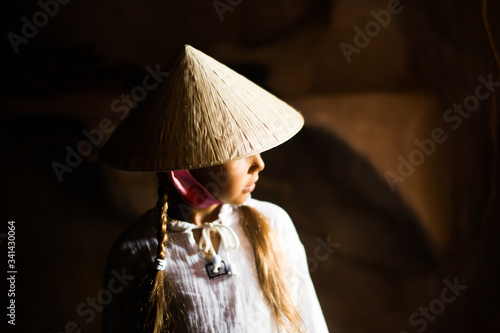 Little girl in Vietnameese hat in dark place with sun light on her face photo