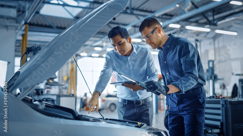 Manager Checks Diagnostics on a Tablet Computer and Explains the Engine Breakdown to a Mechanic. Car Service Employees Inspect Car's Engine Bay with a LED Lamp. Modern Clean Workshop.