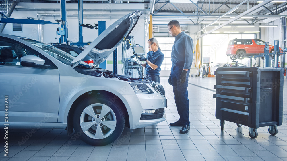 Two Mechanics in a Service are Inspecting a Car After They Got the Diagnostics Results. Female Specialist is Comparing the Data on a Tablet Computer. Repairman is Using a Ratchet to Repair the Faults. Two Mechanics in a Service are Inspecting a Car After They Got the Diagnostics Results. Female Specialist is Comparing the Data on a Tablet Computer. Repairman is Using a Ratchet to Repair the Faults.