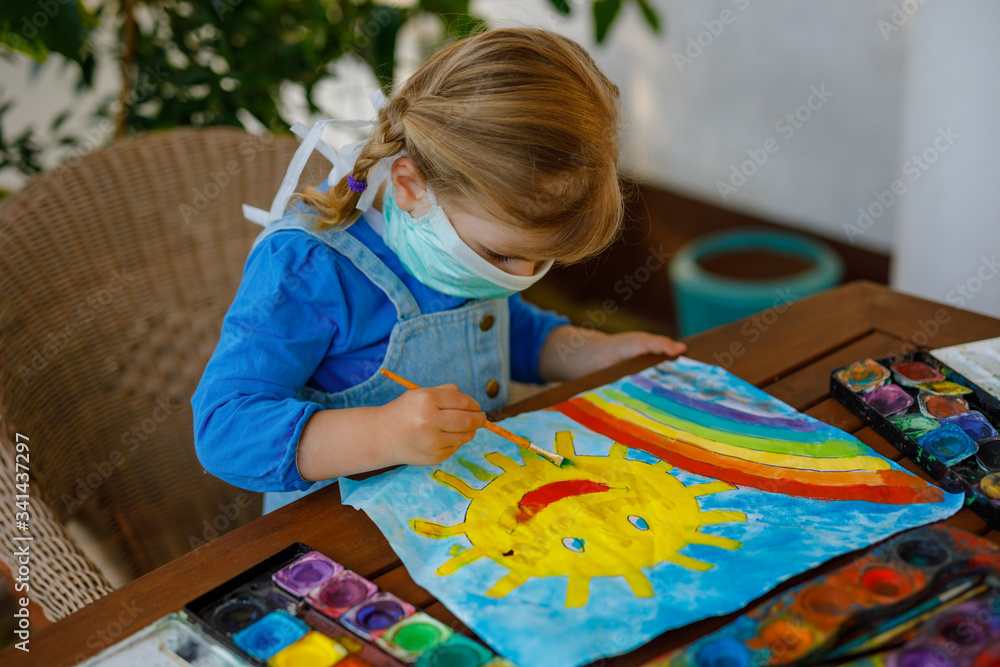 Fototapeta premium little toddler girl in medical mask painting rainbow with water colors during pandemic coronavirus quarantine disease. Children painting rainbows around the world with the words Let's all be well.
