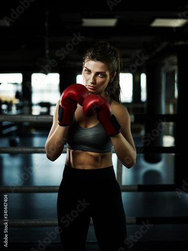 Three quarter length portrait of determined female boxer in red gloves looking confidently at camera while posing in dark ring © Comeback Images
