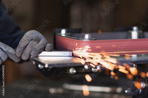 Industrial tool worker grinds a square steel pipe on a rotating belt sander photo