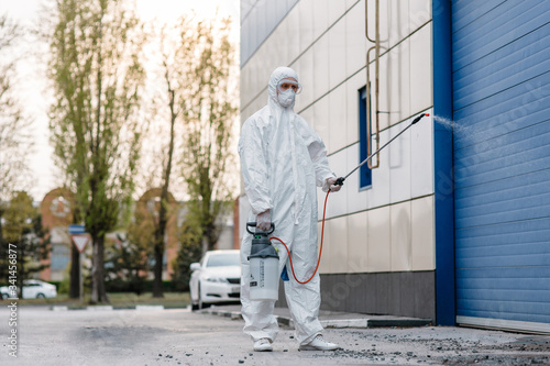 Man wearing an NBC personal protective equipment (ppe) suit, gloves, mask, cleaning the streets with a pressurized spray disinfectant water to remove covid-19 coronavirus. Concept of a pandemic. © Galina Geier