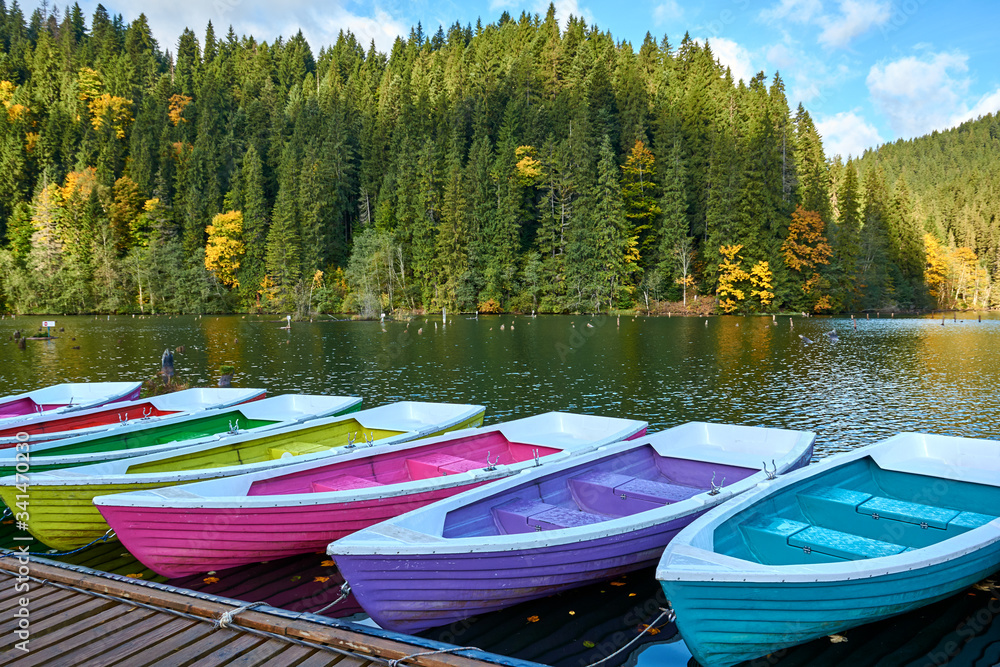 Fototapeta premium Multi-colored boats at the pier on Lake Lacu Rosu, Romania. Autumn landscape of mountain lake, forest and mountains.