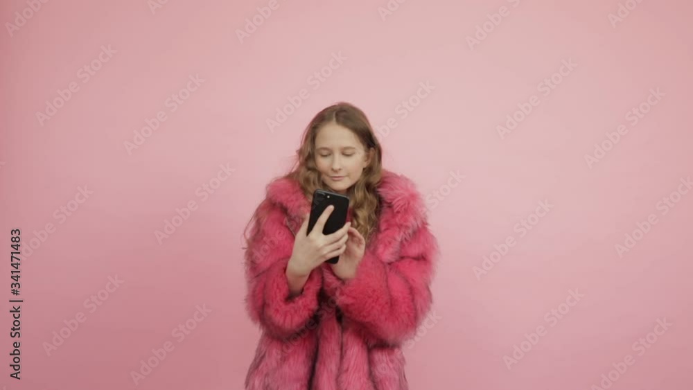 A young girl with long hair in a pink coat and a bright pink dress holds a phone in her hands on a pink background. A young girl with long hair in a pink coat and a bright pink dress holds a phone in her hands on a pink background.
