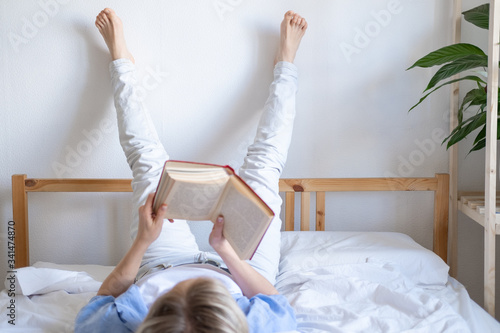 blonde woman holding open book and reading lying on bed legs up on the wall. Leisure, stay home and quarantine. Time for yourself