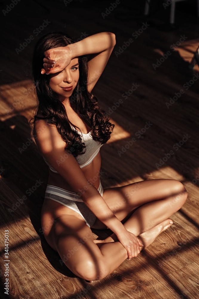 Fototapeta premium Photo of a beautiful female model posing while sitting on a wooden floor in a shade of the window frame wearing sporty underwear