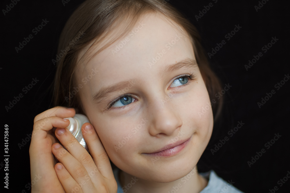 Cute smiling blonde girl holding seashell next to her ear and listening to the ocean on black background in studio Cute smiling blonde girl holding seashell next to her ear and listening to the ocean on black background in studio