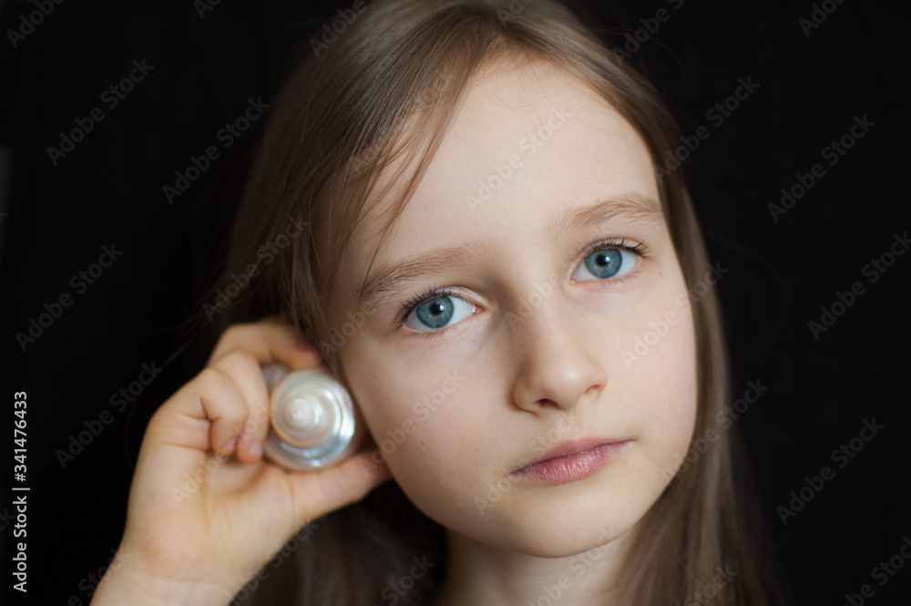 Little girl is dreaming about the summer trip to the ocean and listening to a white shell to hear a sea sound on black background in studio Little girl is dreaming about the summer trip to the ocean and listening to a white shell to hear a sea sound on black background in studio