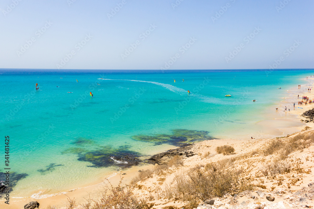 People practice windsurfing on turquoise water in Fuerteventura, Spain. Tourists enjoying leisure activity on sunny day by Sotavento beach. Paradise landscape by the sea in Canary Islands