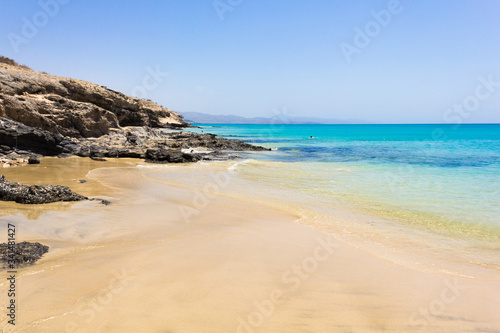 Crystal clear water beach by volcanic rocks coast on sunny day in Fuerteventura. Calm, transparent sea ideal for relax and peaceful vacation. Summer holidays, tourism travel destination concepts