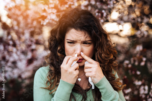 Young woman sneezing in park. She also uses nasal nose drops. Allergy, flu, virus concept