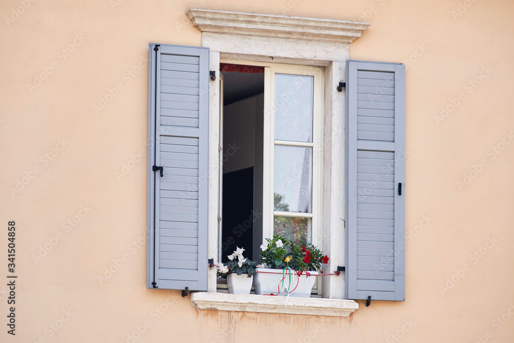Italian window on the pink color wall facade with open wooden grey classic shutters and flowers on the windowsill