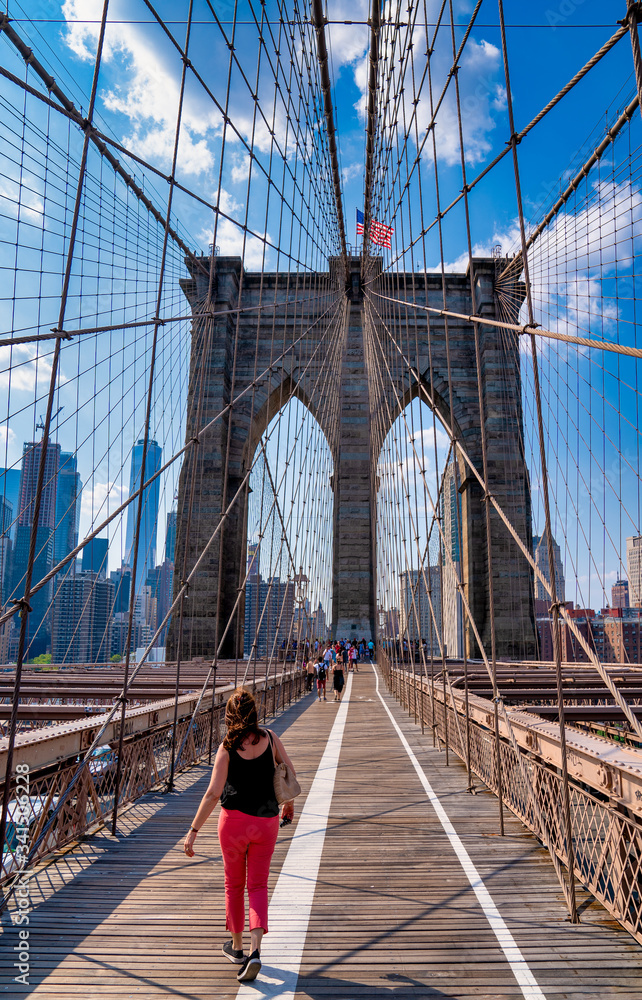 Naklejka premium Brooklin Bridge with American flag atop on a summer day with a woman walking toward the city in red pants