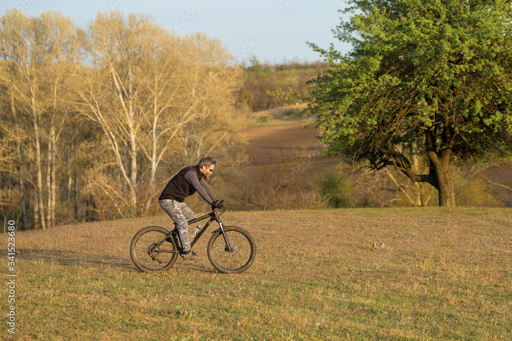 Sports brutal bearded guy on a modern mountain bike. Cyclist on the green hills in the spring.