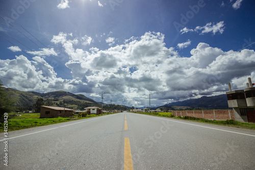 asphalt road that crosses a village with houses, green fields, mountains and gray clouds in the blue sky