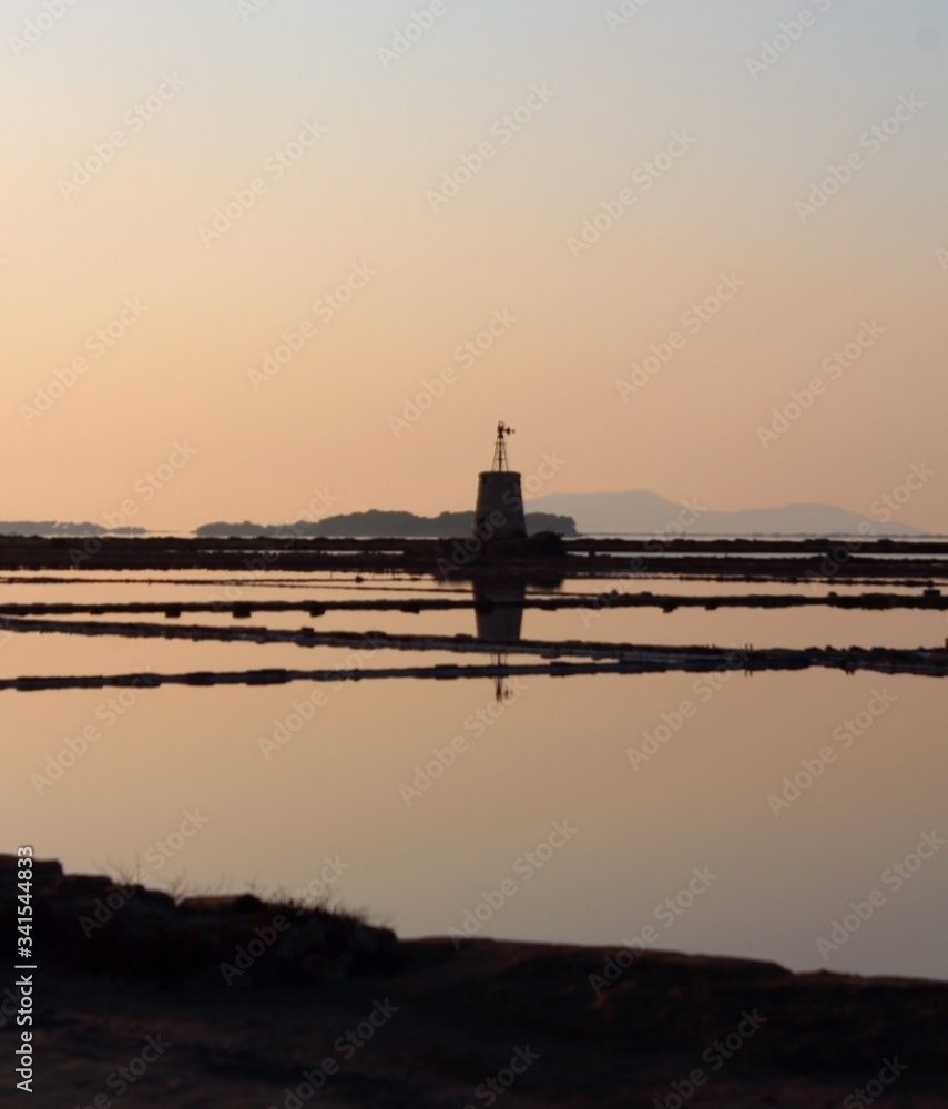 salt pans with water pumping tower and 
promontory in the background at sunset