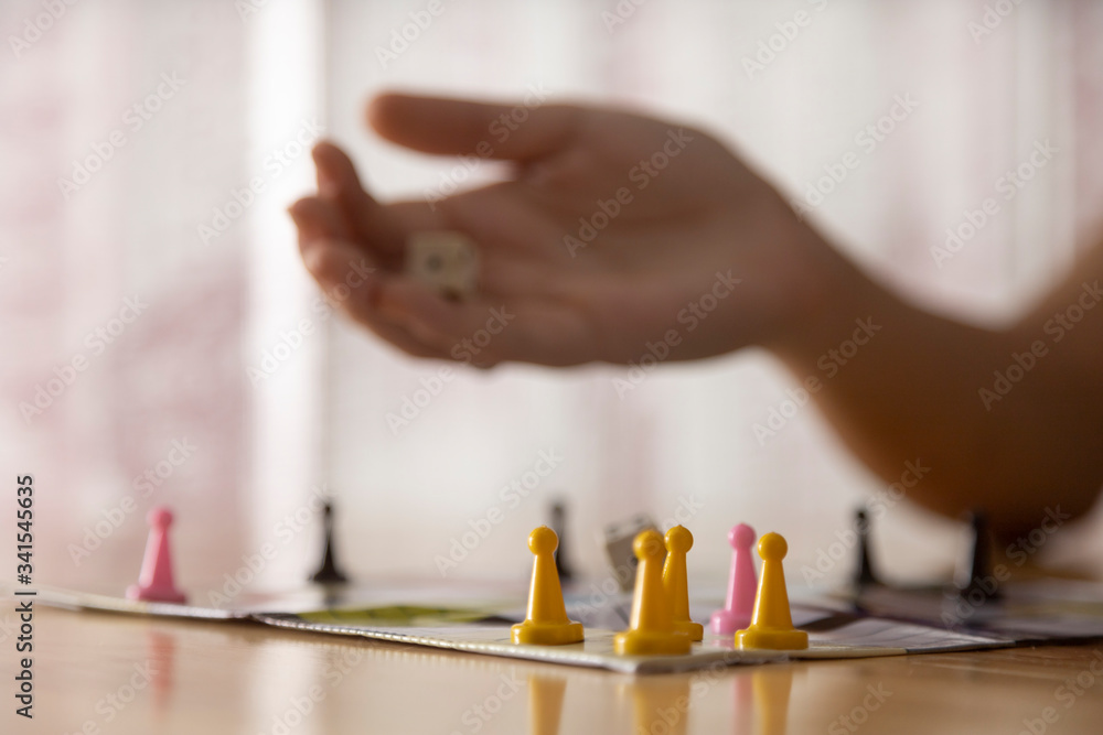 woman's hand throwing board game dice with colored chips on wooden table next to window with curtain and light from outside.