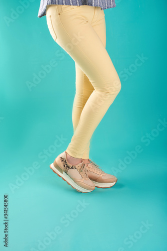 Close up photo of sneakers and yellow skinny jeans against an aqua background in a studio.