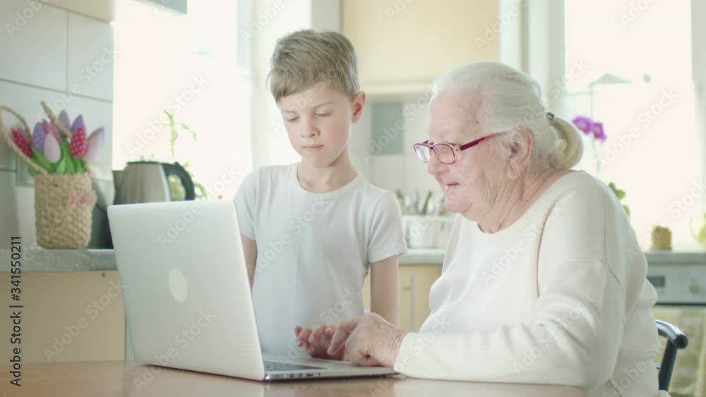 Gray-Haired Grandmother In Glasses Looks Intently At The Laptop Screen. She Is Helped By Little Great-Grandson. Gray-Haired Grandmother In Glasses Looks Intently At The Laptop Screen. She Is Helped By Little Great-Grandson.