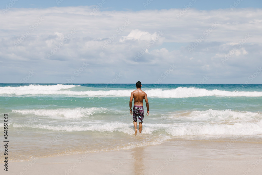 Tan strong man walking in the blue water ocean surrounded by sand on a sunny day Tan strong man walking in the blue water ocean surrounded by sand on a sunny day