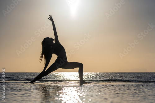 Silhouette of woman standing at yoga pose on the tropical beach during sunset. Girl practicing yoga near sea water
