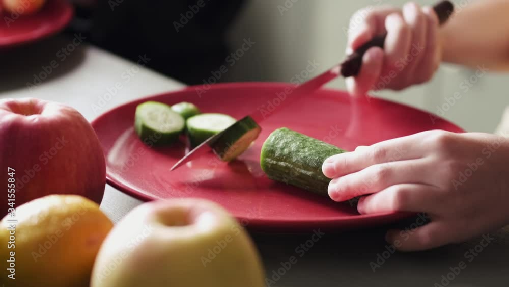 The child is cutting a cucumber on a plate with a knife. The kid is cooking a healthy salads with fresh vegetables. The child is cutting a cucumber on a plate with a knife. The kid is cooking a healthy salads with fresh vegetables.