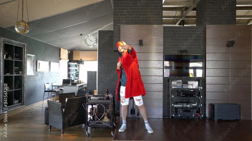 Young unusual guy funny dressed in a red coat with a gold chain around his neck and shorts, he fools around at home in self-isolation and therefore entertains himself and the TikToks audience Young unusual guy funny dressed in a red coat with a gold chain around his neck and shorts, he fools around at home in self-isolation and therefore entertains himself and the TikToks audience