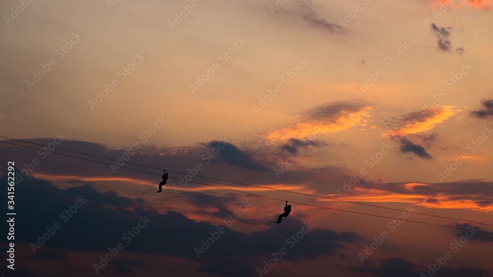 The scenery of the silhouette of the men hanging on the zip line in sunset time.