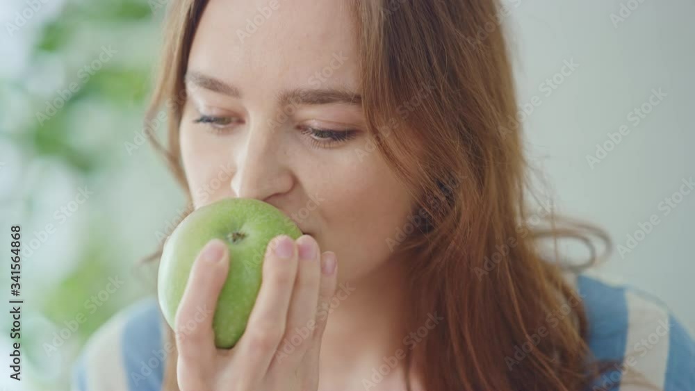 Authentic Portrait of a Beautiful Young Female in a Bright Room at Home Eating an Apple. Attractive Brunette Woman Poses for the Camera and Gently Smiles. Happy Vegetarian Full of Health. Authentic Portrait of a Beautiful Young Female in a Bright Room at Home Eating an Apple. Attractive Brunette Woman Poses for the Camera and Gently Smiles. Happy Vegetarian Full of Health.
