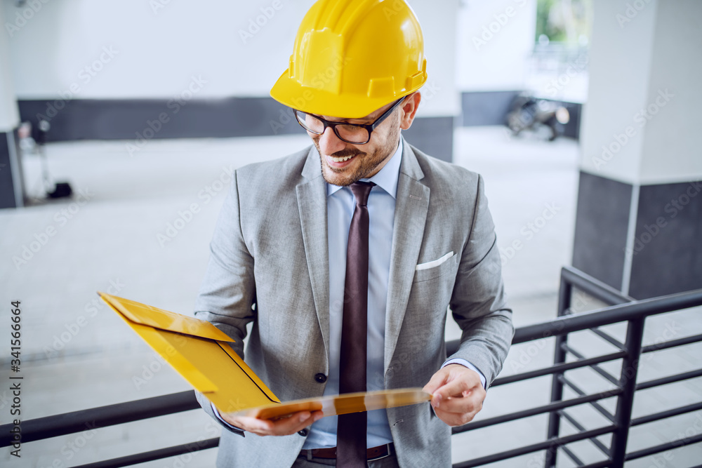 Attractive caucasian elegant architect in suit and with helmet on head leaning on railing and looking at documents in folder.
