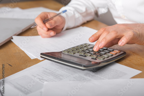 Close up of businessman or accountant hand holding pen working on calculator accountancy document and laptop computer at office  business concept