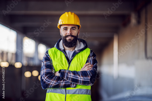 Obraz na plátně Cute Caucasian bearded construction worker with safety helmet on head in vest standing with arms crossed at construction site and looking at camera