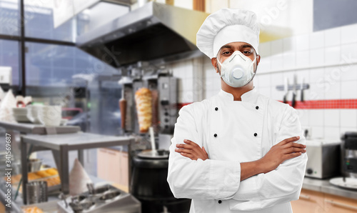 health protection, safety and pandemic concept - indian male chef cook wearing face protective mask or respirator with crossed arms over kebab shop kitchen background