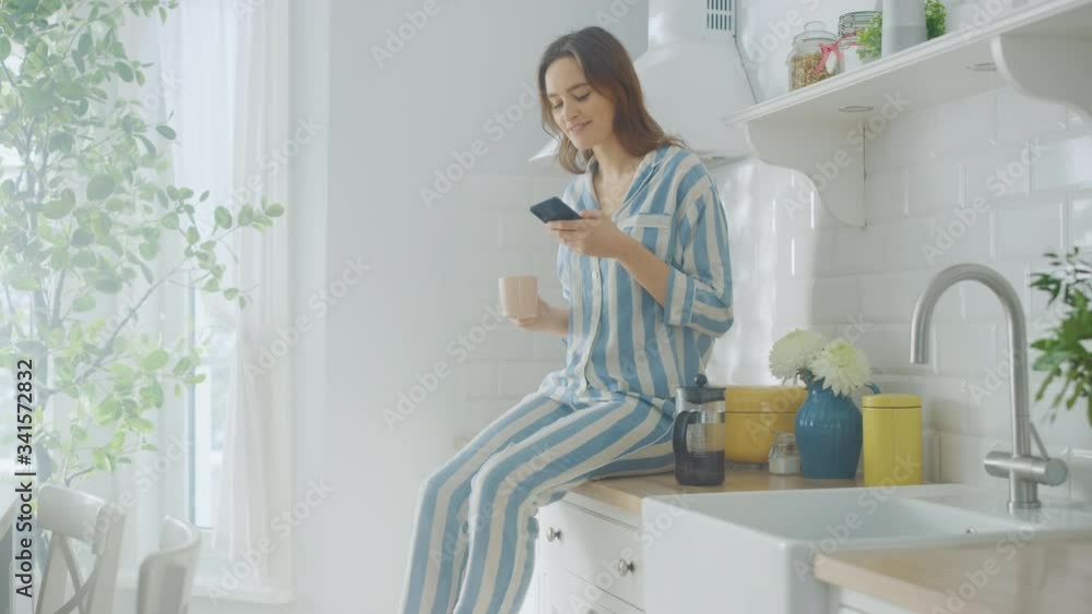 Beautiful Female is Sitting on a Cupboard and Using a Smartphone in a Kitchen While Drinking a Cup of Freshly Brewed Coffee. Girl in Pyjamas with Healthy Lifestyle Relaxes at Home in the Morning. Beautiful Female is Sitting on a Cupboard and Using a Smartphone in a Kitchen While Drinking a Cup of Freshly Brewed Coffee. Girl in Pyjamas with Healthy Lifestyle Relaxes at Home in the Morning.