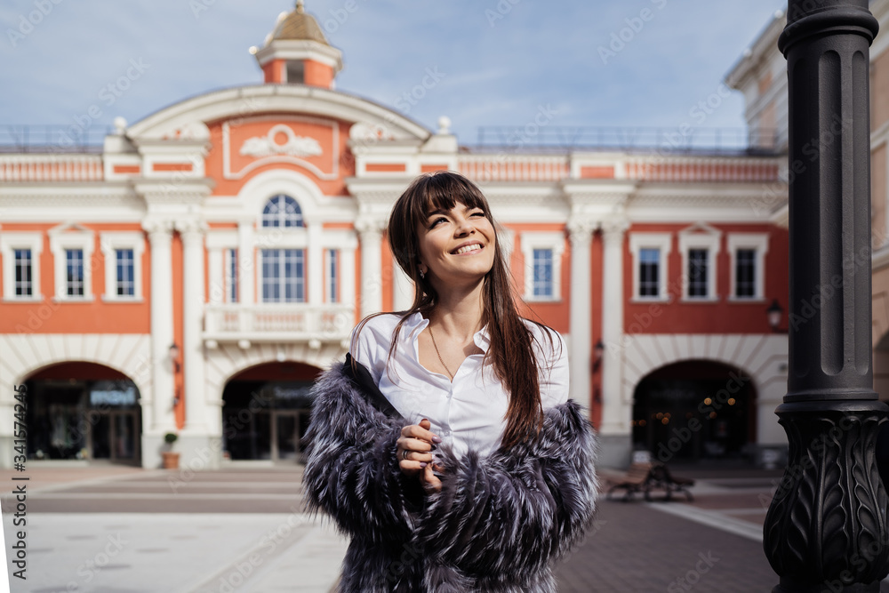 beautiful smiling brunette with the long straight hair wearing in a fur coat and the white skirt standing against the background of  the classic style building smiles looking at the sunny blue sky