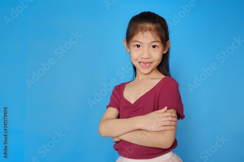 An attractive and confident preteen Asian girl with her arms crossed, smiling. Plain light blue background.