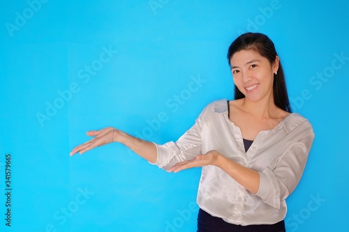 A waist height shot of an attractive Asian woman in her 30s presenting a product, pointing with open hands on a light blue plain background.