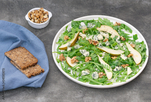 Vegetarian salad with arugula leaves, pear, blue cheese, and walnuts. Some dark bread on blue napkin and more walnuts on the table. Healthy lunch or dinner. 