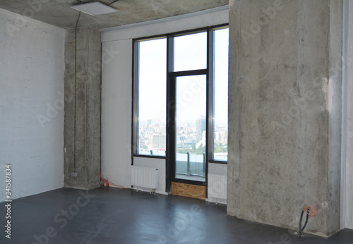 Room renovation in the apartment building with a window and a balcony under construction, concrete floor, pillar in the corner and walls without plastering and finishing.