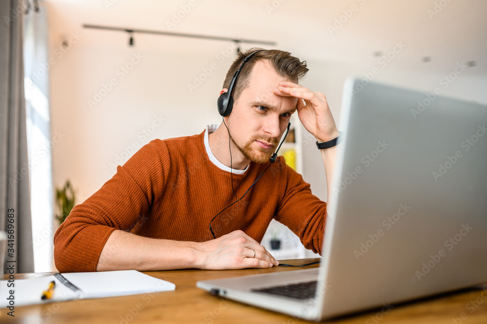 A guy call center operator, support service operator at home office. He uses a headset to talk sits with a cup of coffee at a table, looks at a laptop screen with serious face A guy call center operator, support service operator at home office. He uses a headset to talk sits with a cup of coffee at a table, looks at a laptop screen with serious face
