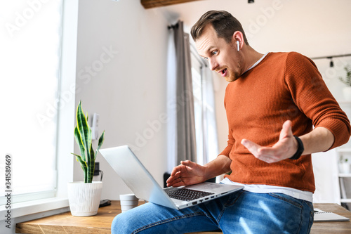 Unexpected turn. A young bearded guy with a laptop indoors. He sits on the edge of table, looks at the screen with suprised face photo