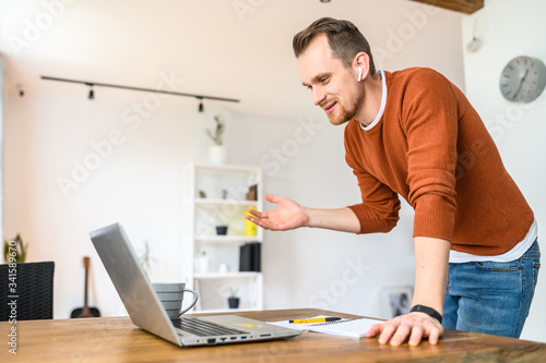 Video chat online, video call, zoom. A young bearded guy speaks using a laptop. He is standing, looking at the camera, explaining something and smiling.