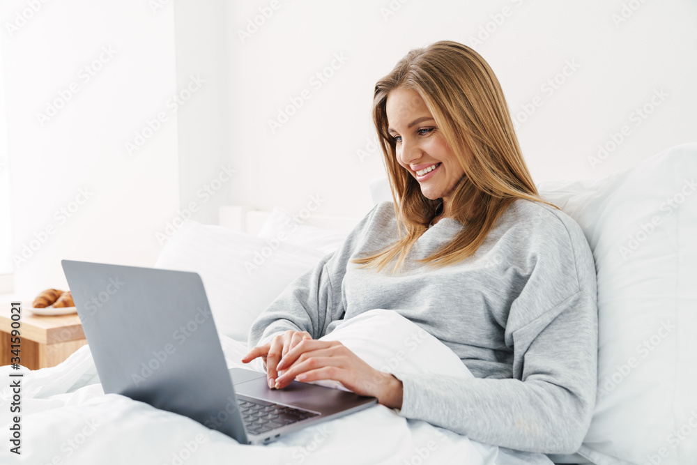 Image of woman smiling and using laptop while lying in bad Image of woman smiling and using laptop while lying in bad