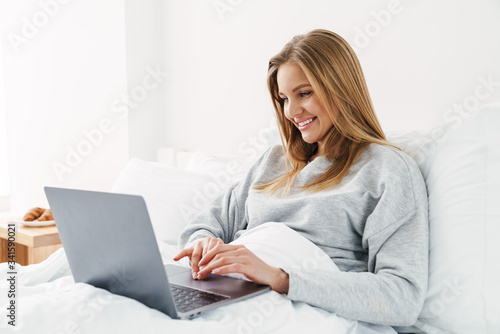 Image of woman smiling and using laptop while lying in bad © Drobot Dean