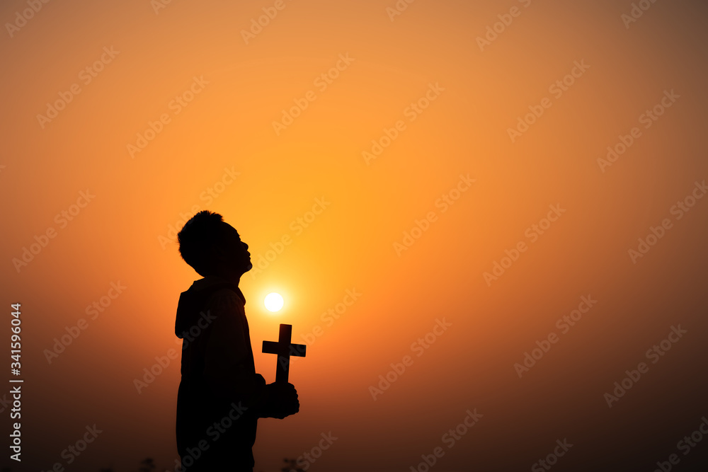 Boy praying to God with the cross in hands on the mountain with light sunset background, Christians should worship and thank God, Christian silhouette concept.